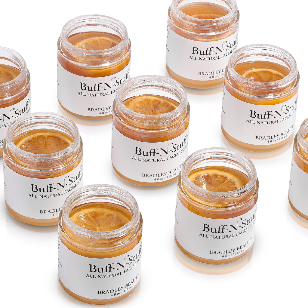 Artistic photo of Buff-N-Stuff All-Natural Facial Scrub with multiple product jars shown without lids. A lemon slice can be seen floating on top of golden scrub product inside each jar