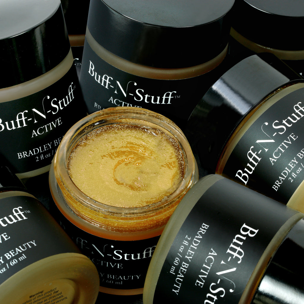 Artistic photo of Buff-N-Stuff Active Facial Scrub showing multiple jars of product laying about and up against each other with a single open jar in the center with cap removed showing the grainy gold texture of the product 